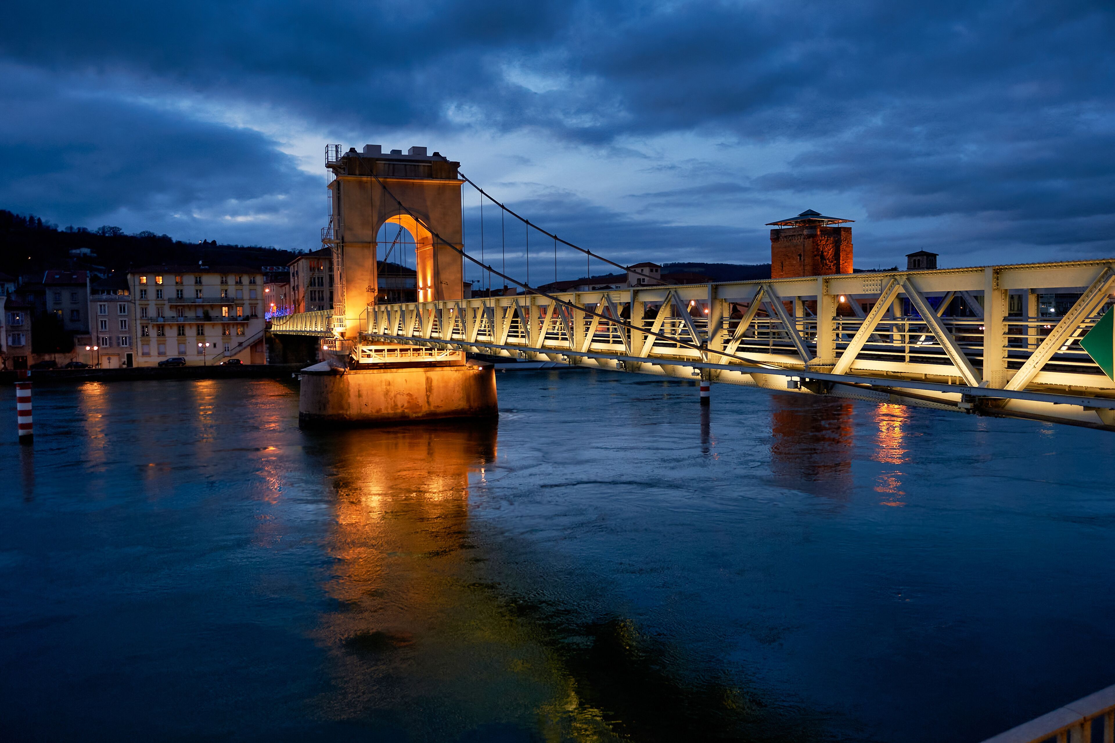 A bridge with a central arch and a tower, spanning over a body of water with buildings in the background.