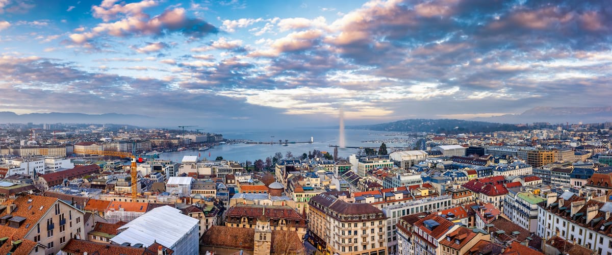 A panoramic view of a city with a river, a bridge, and a distant mountain range under a partly cloudy sky.