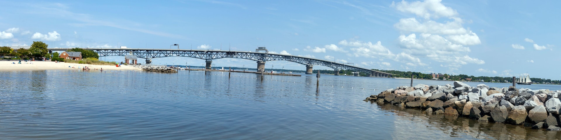 A steel bridge over a calm river with a sandy beach and rocky outcrops on the right.