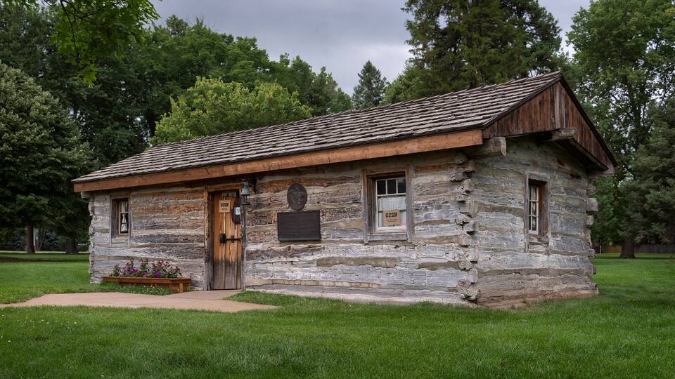 A rustic log cabin with a wooden door and windows, surrounded by lush green grass and trees.