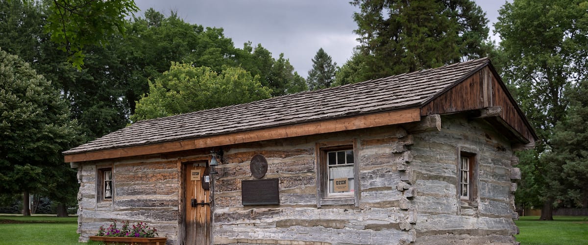 A rustic log cabin with a wooden door and windows, surrounded by lush green grass and trees.