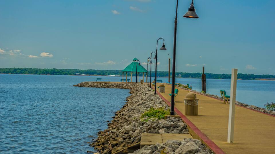 A waterfront promenade with a gazebo, benches, and street lamps.