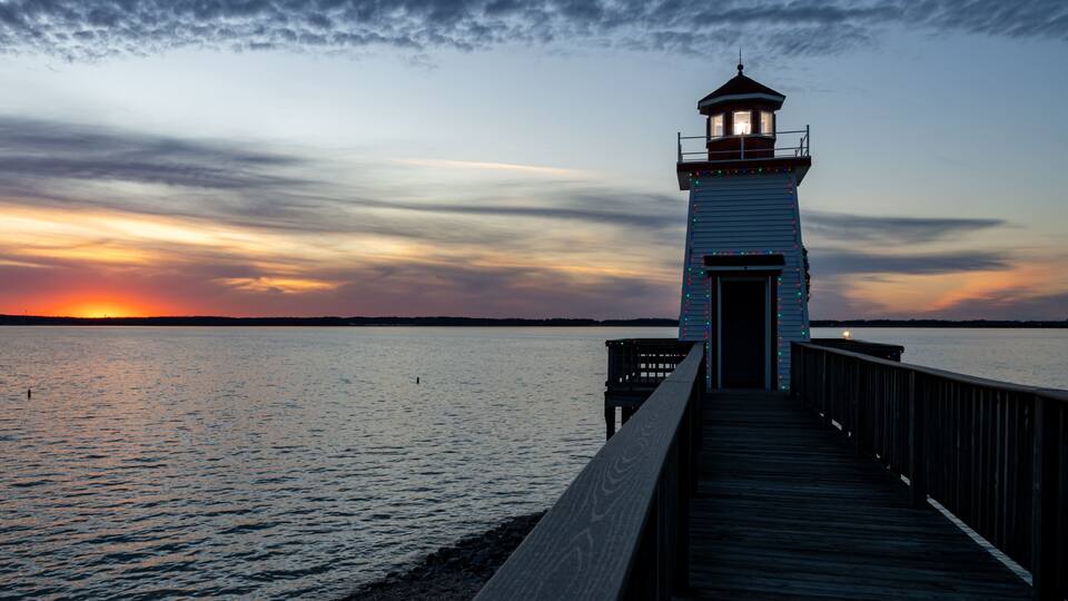A lighthouse at sunset with a wooden pier leading to it.