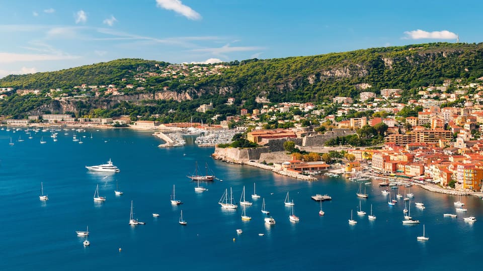 A coastal town with a harbor filled with boats, a castle on a hill, and buildings along the waterfront.
