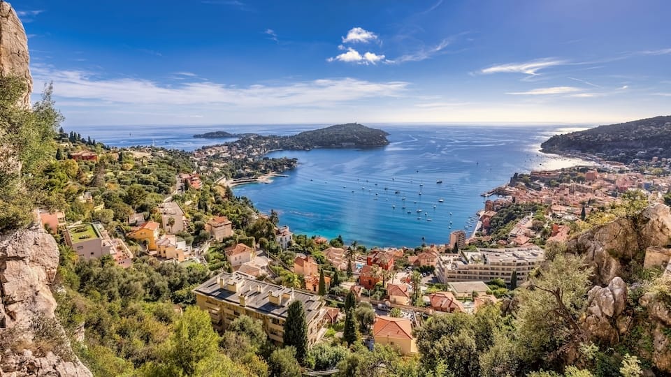 A coastal town with a clear blue sea, rocky terrain, and dense greenery.