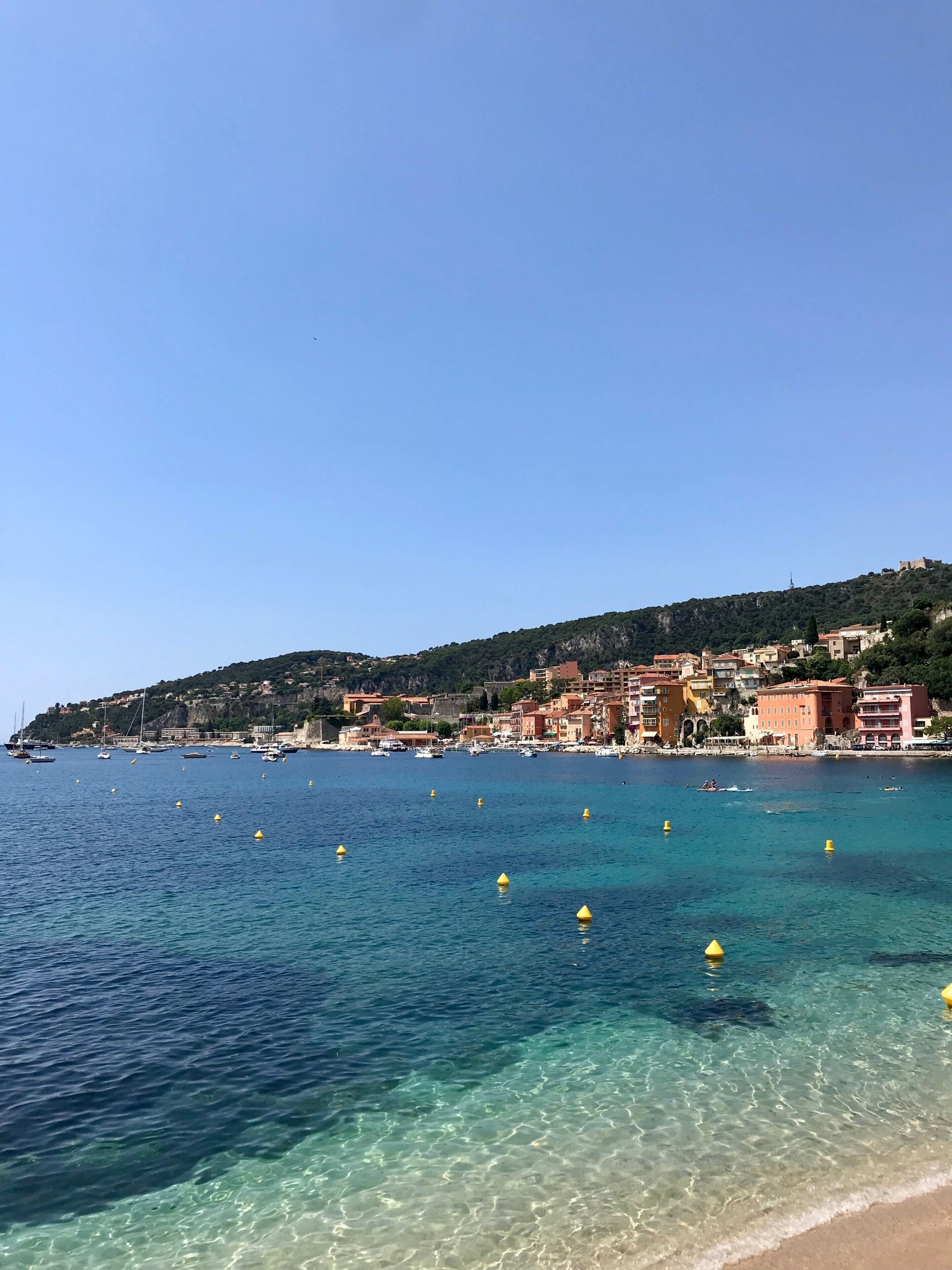 A coastal town with clear blue waters, yellow buoys, and historic buildings on a hillside.