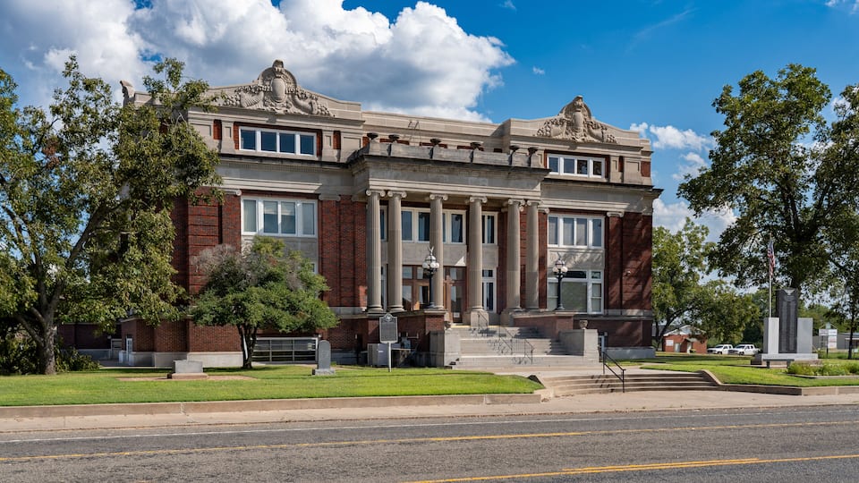 A historic building with classical architecture, including columns and decorative elements, surrounded by a well-maintained lawn and trees.