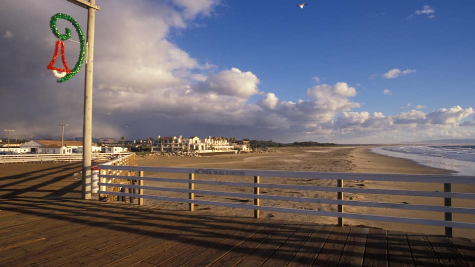 A wooden boardwalk leading to a beach with a decorative sign, a cloudy sky, and buildings in the distance.
