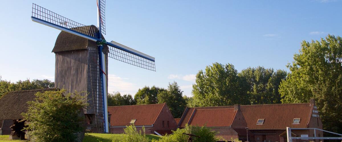 A traditional windmill with a wooden structure and a metal windlass, surrounded by houses with red-tiled roofs and lush greenery.