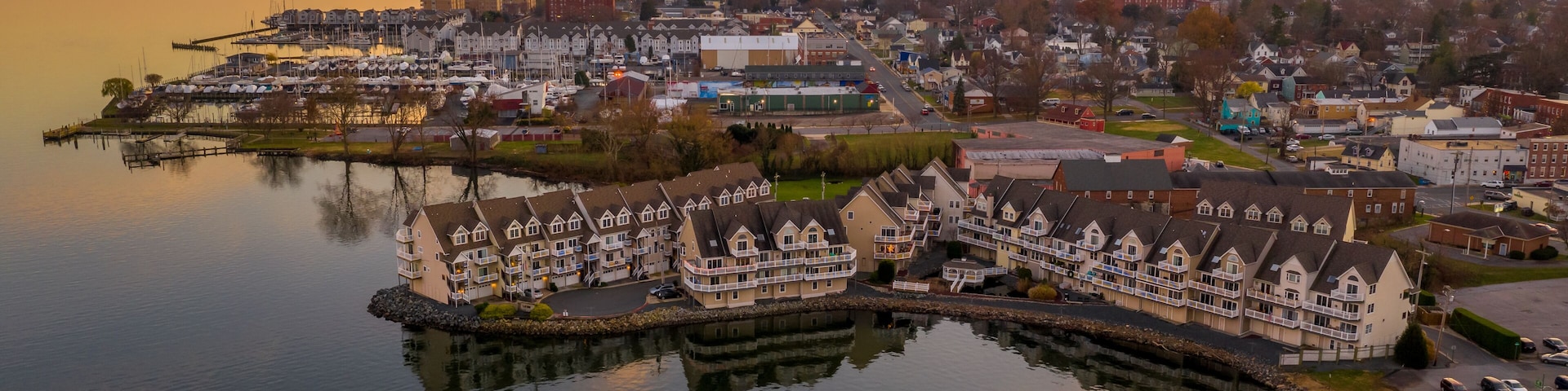 Un pueblo costero con un puerto deportivo, casas frente al mar y un atardecer sobre el agua.