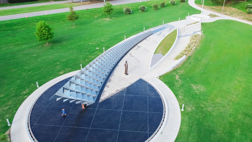 A modern bridge with a unique geometric design over a body of water, surrounded by green lawns and trees.