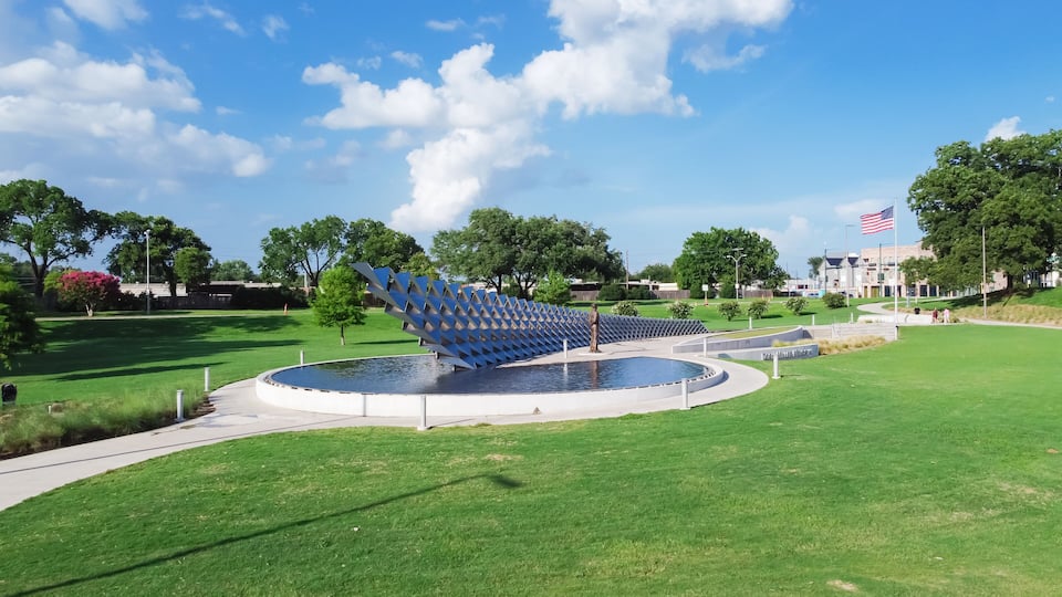 A sculpture of a woman in a blue dress in a park with a circular water feature and a flag.