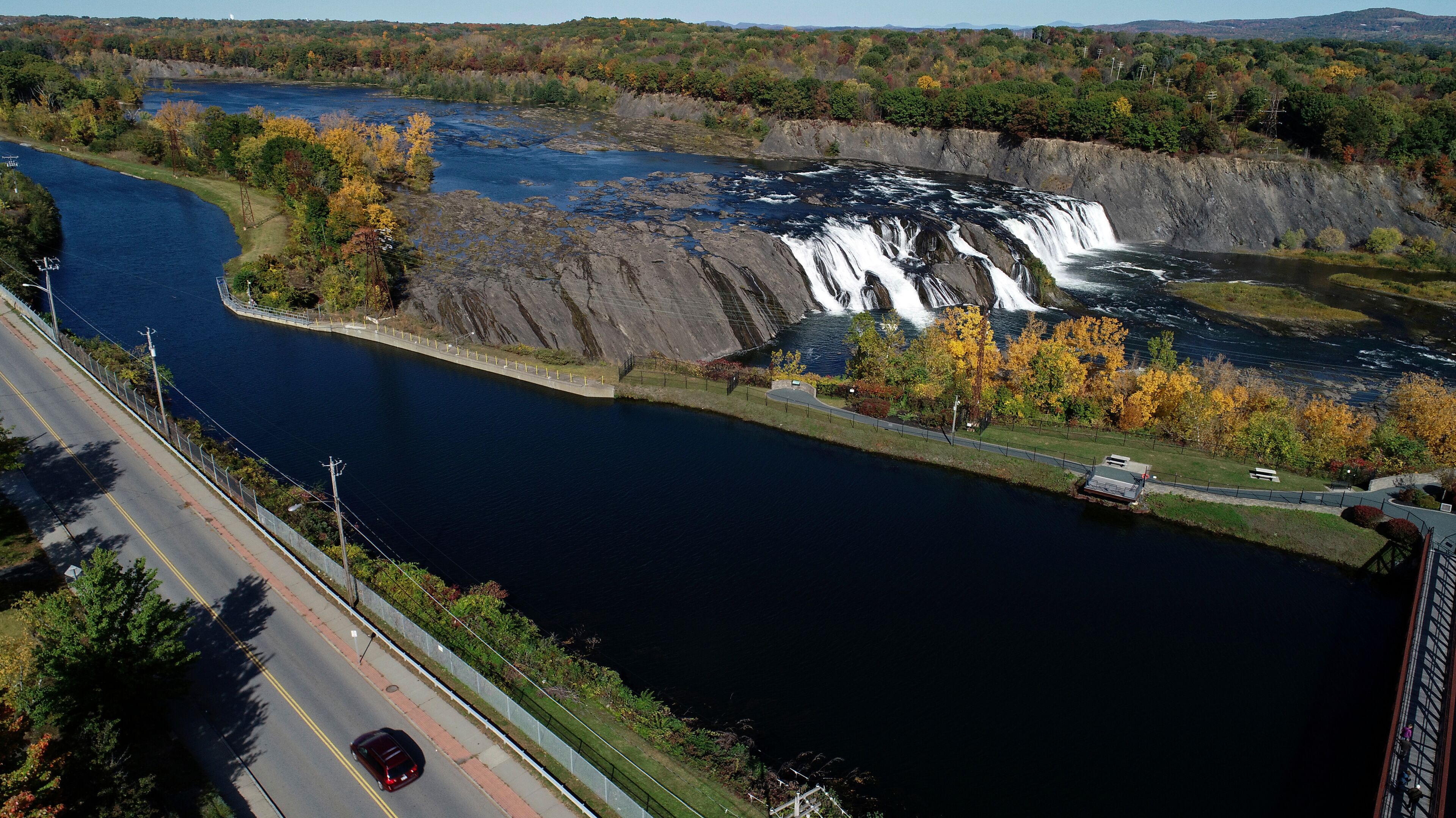 A river with a waterfall, a road, and autumn foliage.