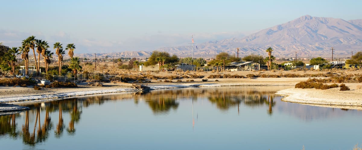 A calm river with reflections, palm trees, a mountain, and sparse buildings in the background.