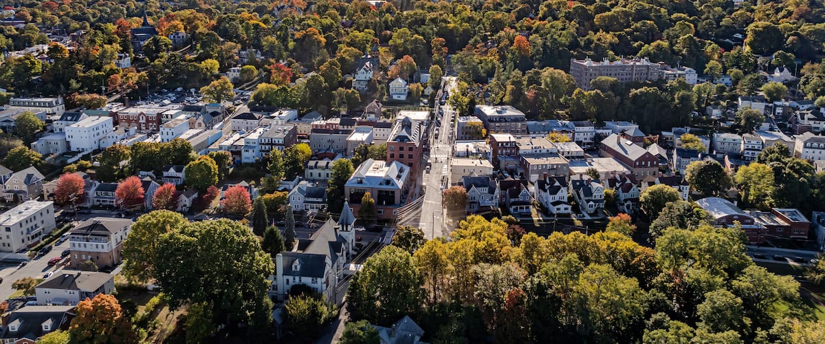 An aerial view of a town with a central street, surrounded by autumn-colored trees and residential buildings.