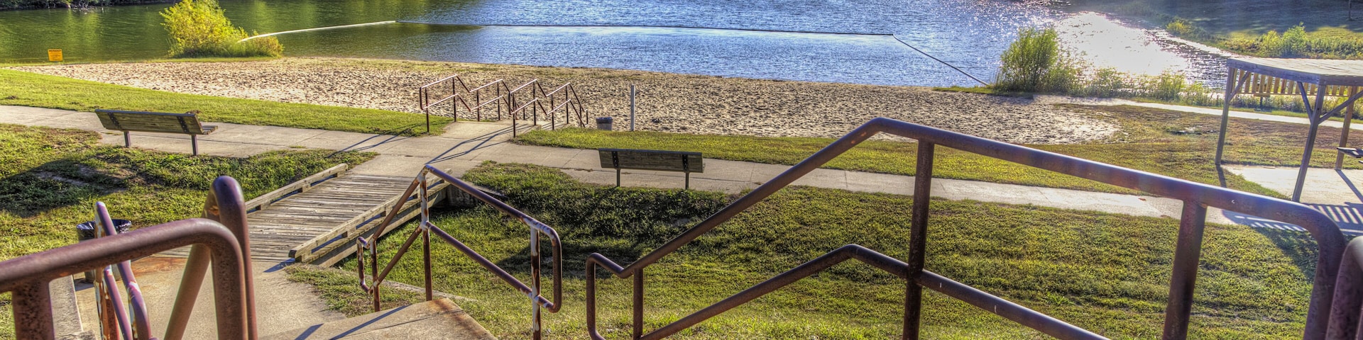 Un parque junto al lago con una escalera de hormigón, barandillas metálicas y mesas de picnic.