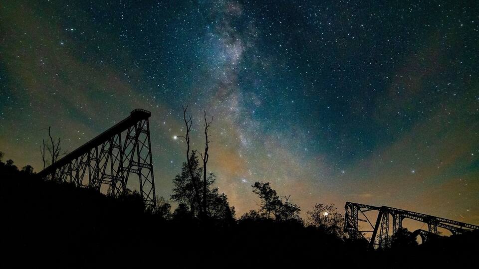 A night sky with stars and the Milky Way, a bridge, and trees silhouetted against the sky.