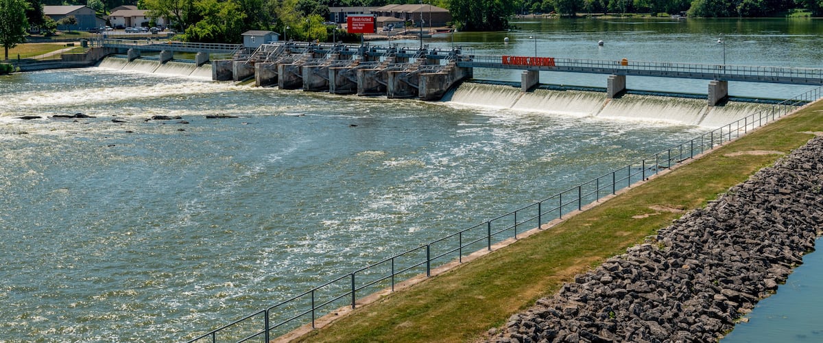 Un barrage avec de l’eau qui s’écoule par-dessus, un pont et une rivière bordée de verdure.