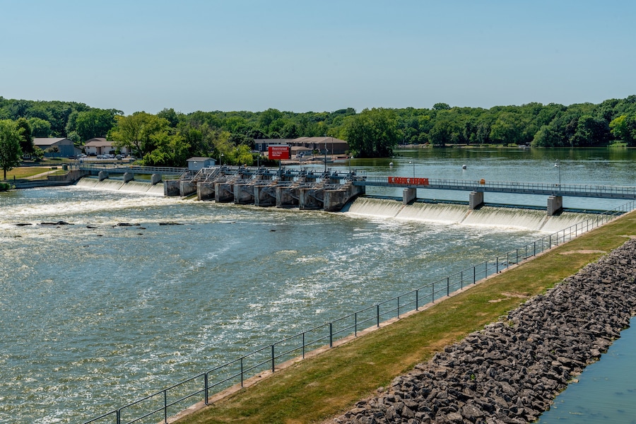 Una diga con acqua che scorre, un ponte e un fiume circondato da vegetazione.
