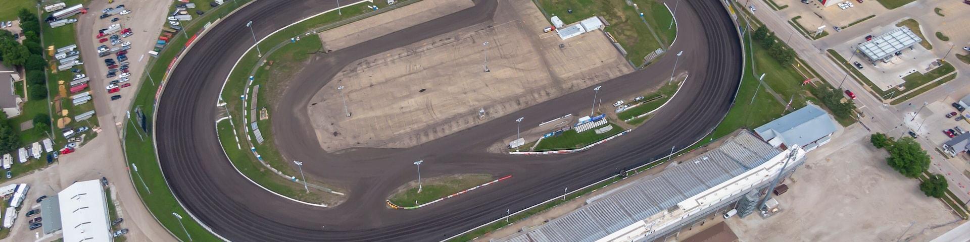 An aerial view of a racetrack with a grandstand, parking areas, and surrounding buildings.