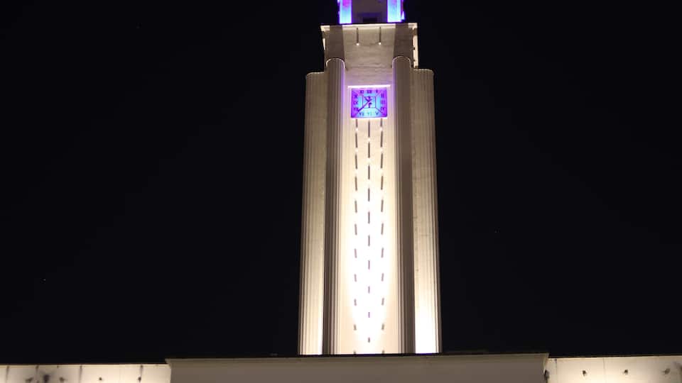 A tall tower with a clock face illuminated in purple at the top.