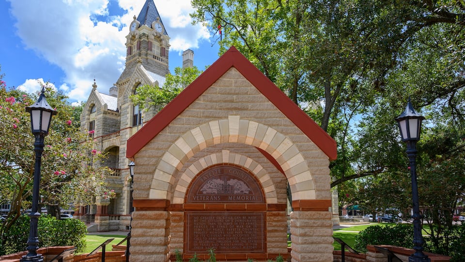 A stone Veterans Memorial with an arched entrance, a clock tower in the background, and surrounding greenery.