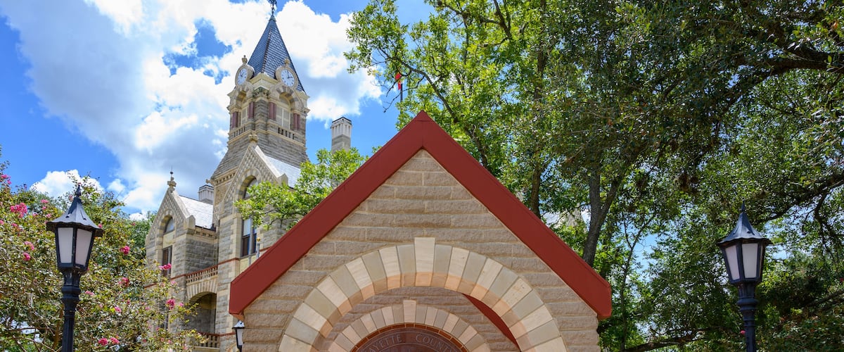 A stone Veterans Memorial with an arched entrance, a clock tower in the background, and surrounding greenery.