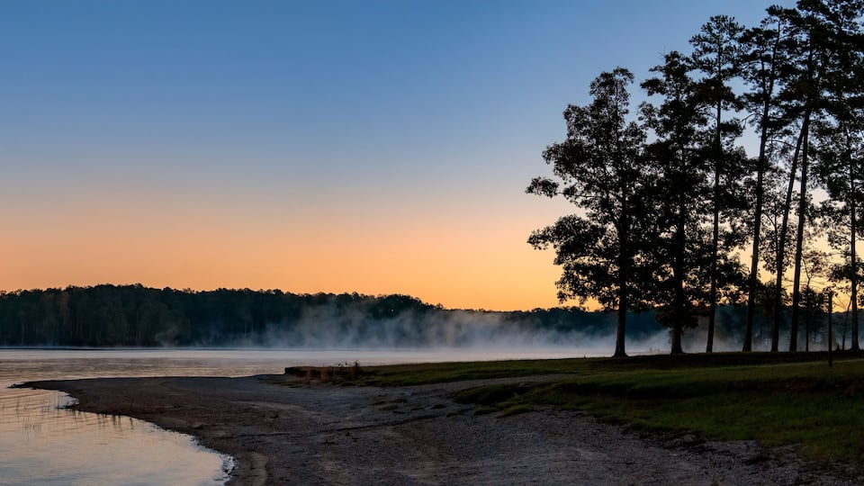 A calm lake at sunset with a forested shoreline and a mist rising from the water.