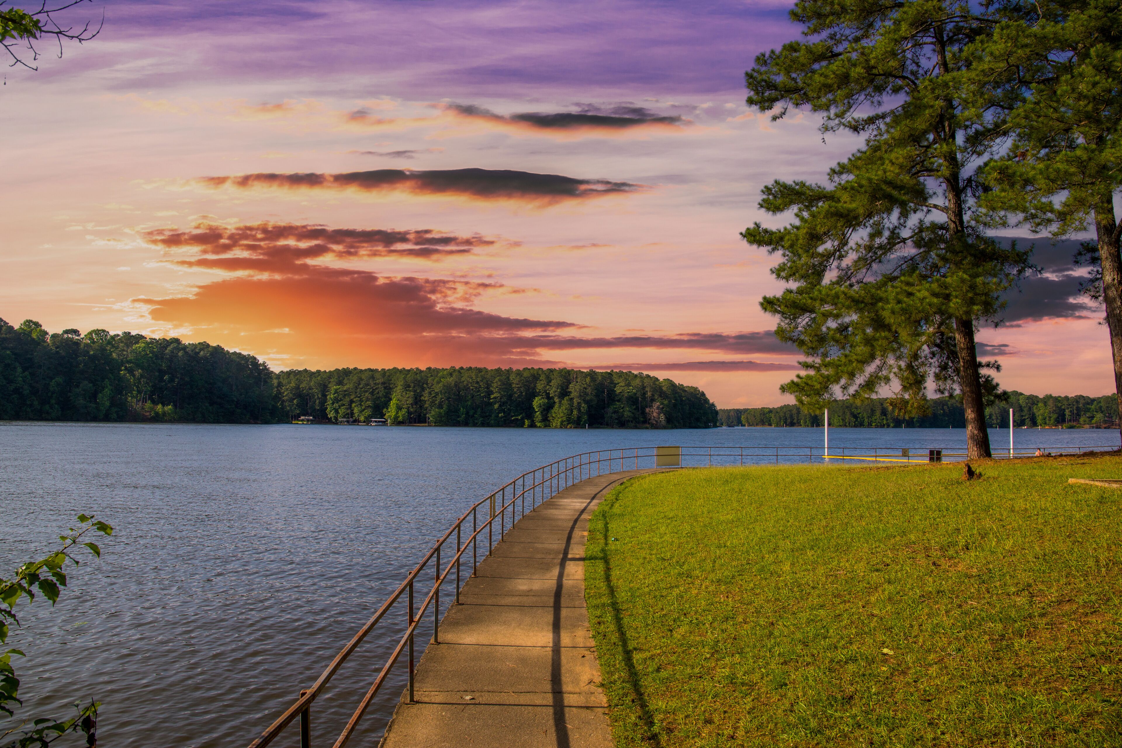 the gorgeous blue rippling waters of Lake Acworth with a long winding footpath around the lake surrounded by a metal rail and lush green trees, grass and plants and powerful clouds at sunset