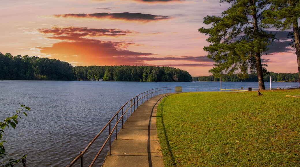 the gorgeous blue rippling waters of Lake Acworth with a long winding footpath around the lake surrounded by a metal rail and lush green trees, grass and plants and powerful clouds at sunset