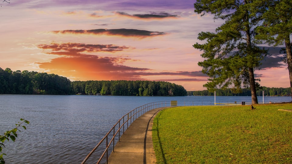 A lakeside path with a railing, a grassy area, and a forested shoreline.