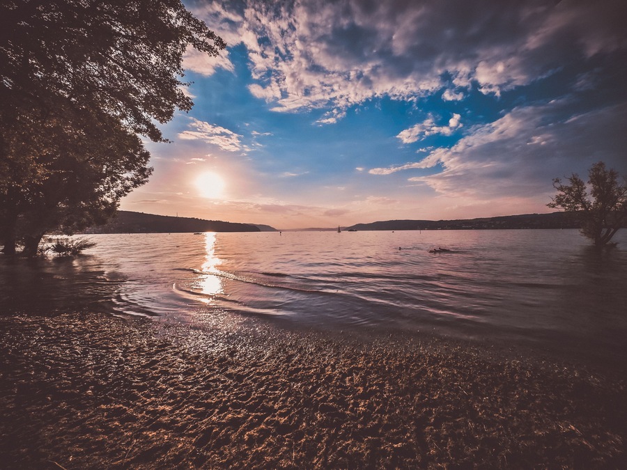 Sunshine over a calm lake with reflecting water and a vast sky. The sandy beach in the foreground invites relaxation.