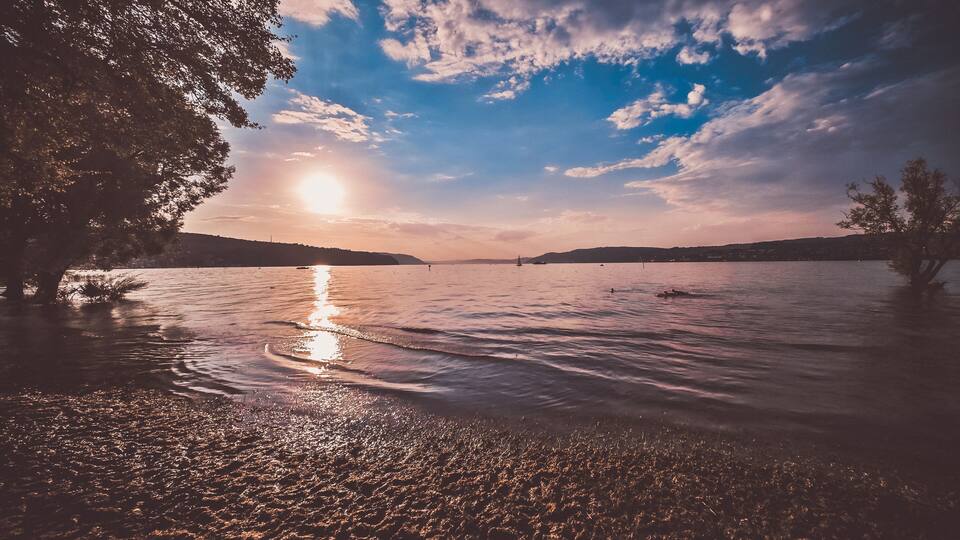 Sunshine over a calm lake with reflecting water and a vast sky. The sandy beach in the foreground invites relaxation.