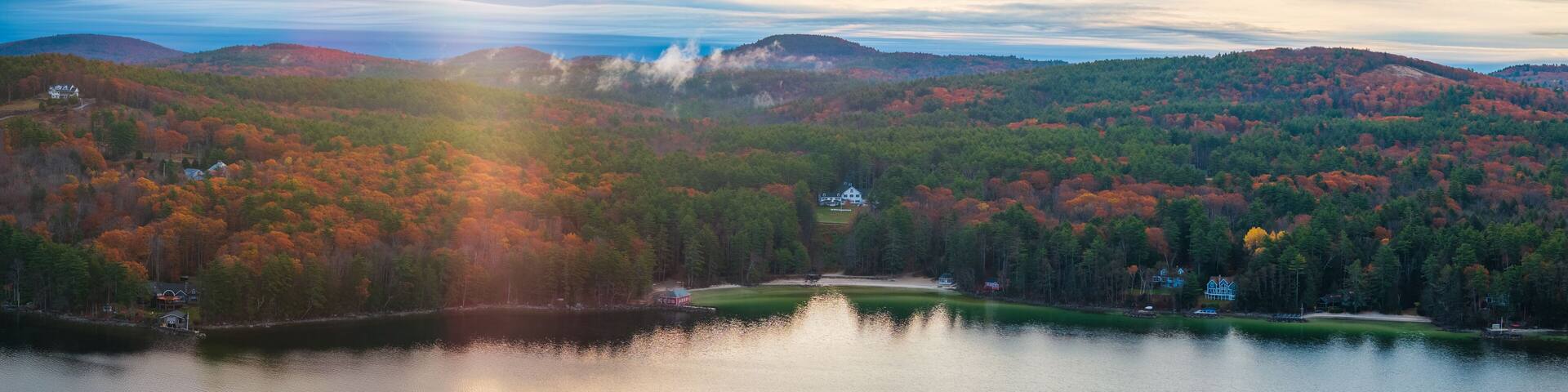 Aerial view of a serene lake reflecting the vibrant autumn foliage under a hazy sky, Alton, New Hampshire, United States.