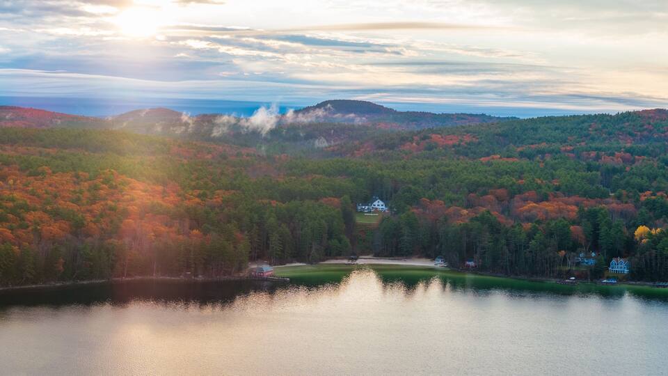 Aerial view of a serene lake reflecting the vibrant autumn foliage under a hazy sky, Alton, New Hampshire, United States.