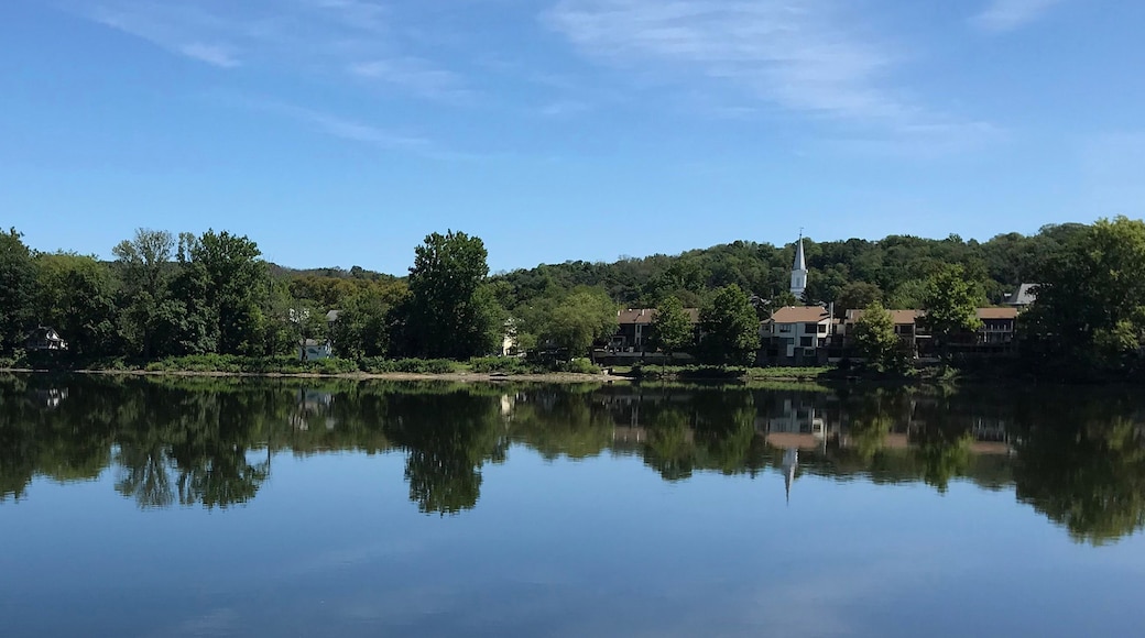 Ein stiller See mit Spiegelungen von Häusern und Bäumen, umgeben von üppigem Grün und einem klaren Himmel.