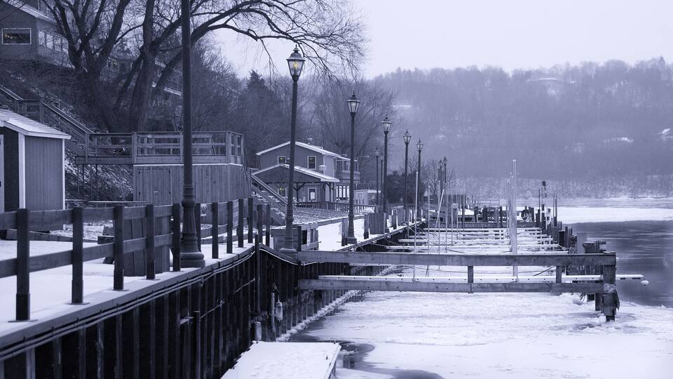 A snow-covered pier extending into a frozen body of water, flanked by buildings and bare trees.