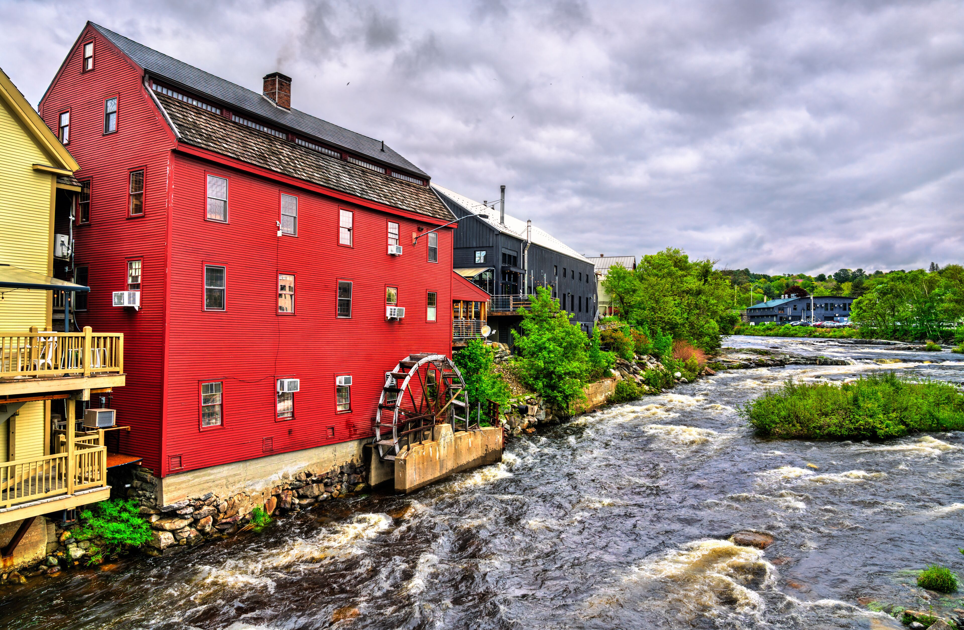 In Littleton, New Hampshire, United States, a red clapboard grist mill with a working waterwheel stands above the rushing Ammonoosuc River beneath a bright, cloud-dappled summer sky.