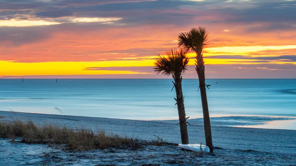 A beach with two palm trees and a sunset over the ocean.