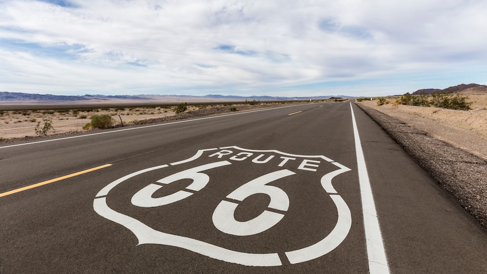 A road with Route 66 markings, surrounded by desert landscape and mountains.