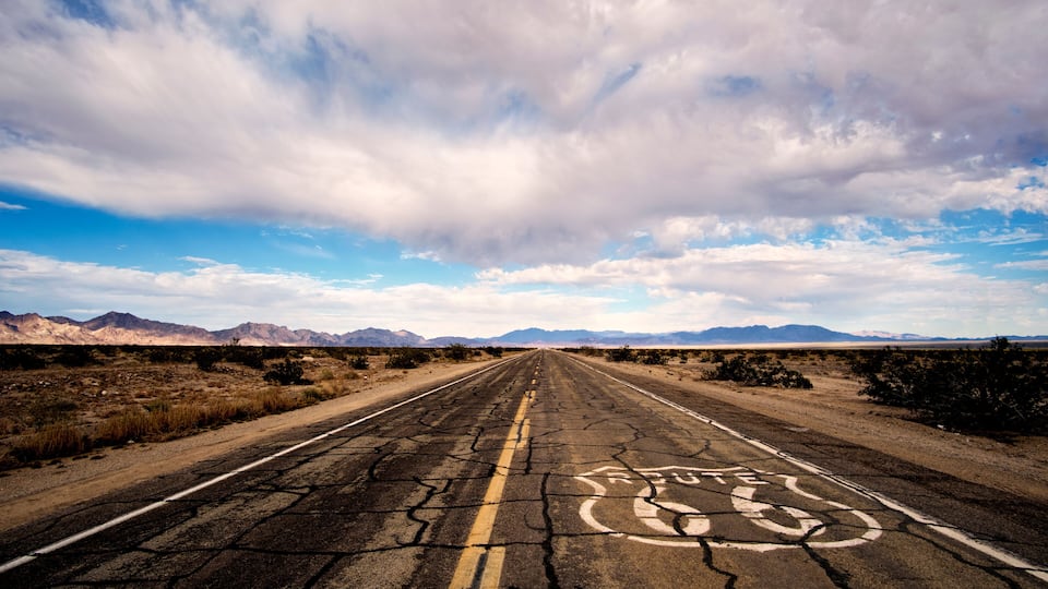 A cracked road with a circular symbol in the center, surrounded by a desert landscape with mountains in the distance under a partly cloudy sky.