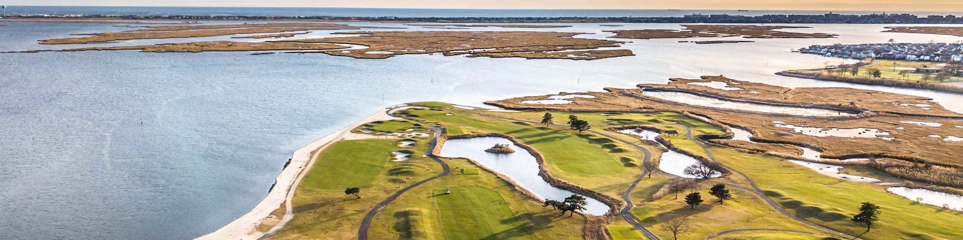 An aerial view of a coastal golf course with water bodies and a distant town.