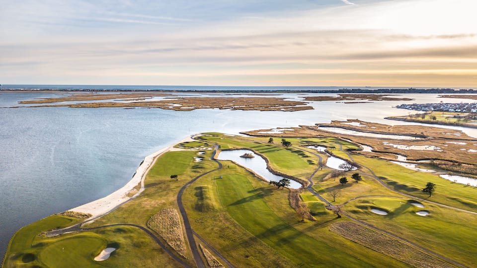 An aerial view of a coastal golf course with water bodies and a distant town.