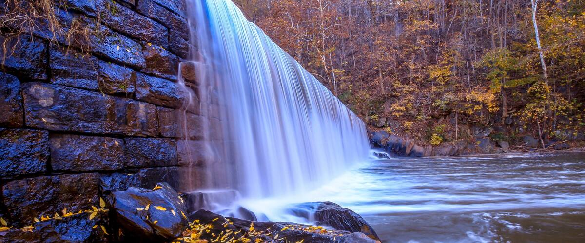 A waterfall cascading over dark rocks, surrounded by autumn foliage.