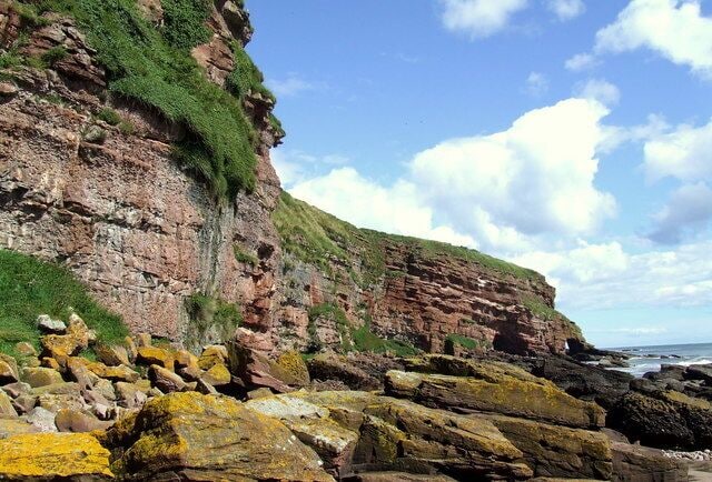 Boulders under cliffs, Auchmithie The boulders under the cliff at this point are above the high water point and as such have a thick covering of yellow lichen. In the background is a headland with an arch. On the OS map this is marked as Maw Skelly I am unsure if this refers to the headland and if the arch has a name of its own?