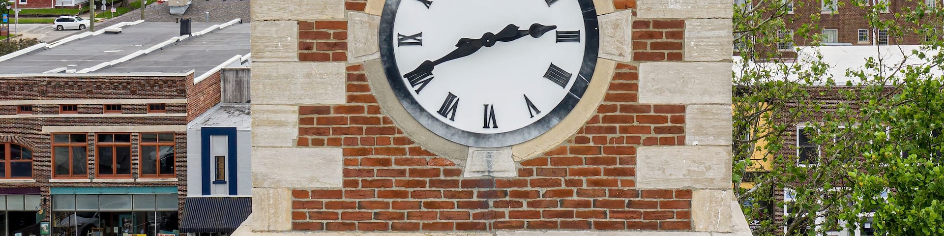 A clock tower with Roman numerals and a white face, set against a brick wall with a metal roof.