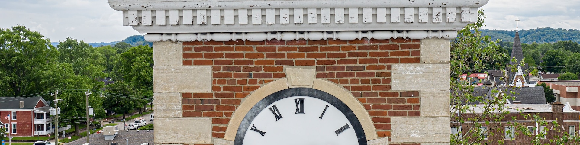 A clock tower with Roman numerals and a white face, set against a brick wall with a metal roof.