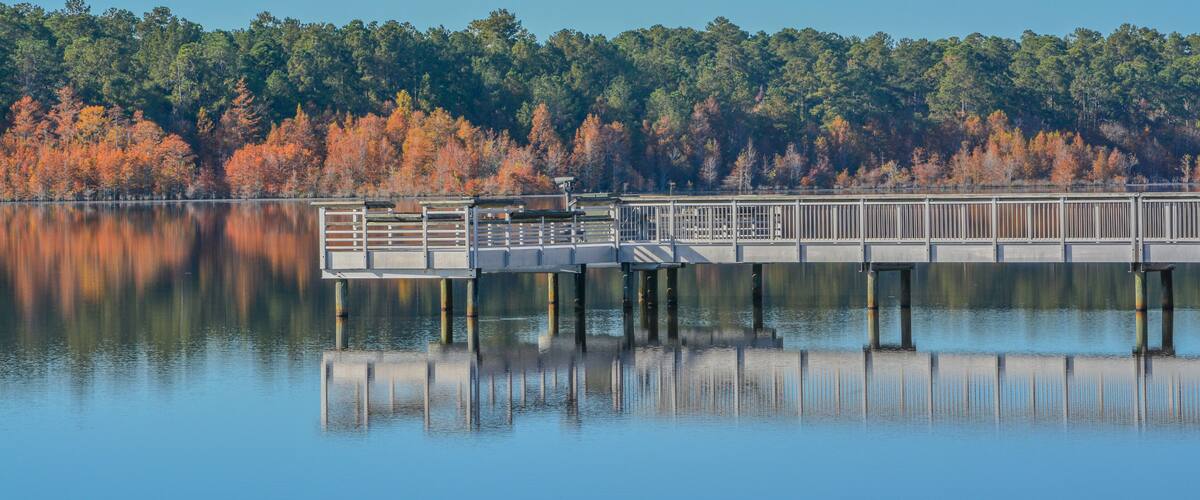 A calm lake with a wooden pier extending into the water, surrounded by autumn-colored trees.
