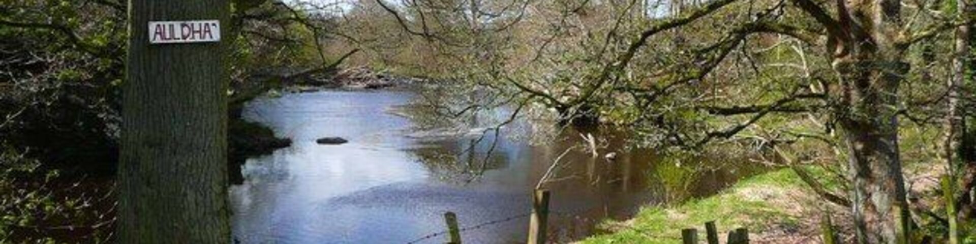 Un río tranquilo con un cartel que dice "ALLDHA" y un área cercada con árboles.