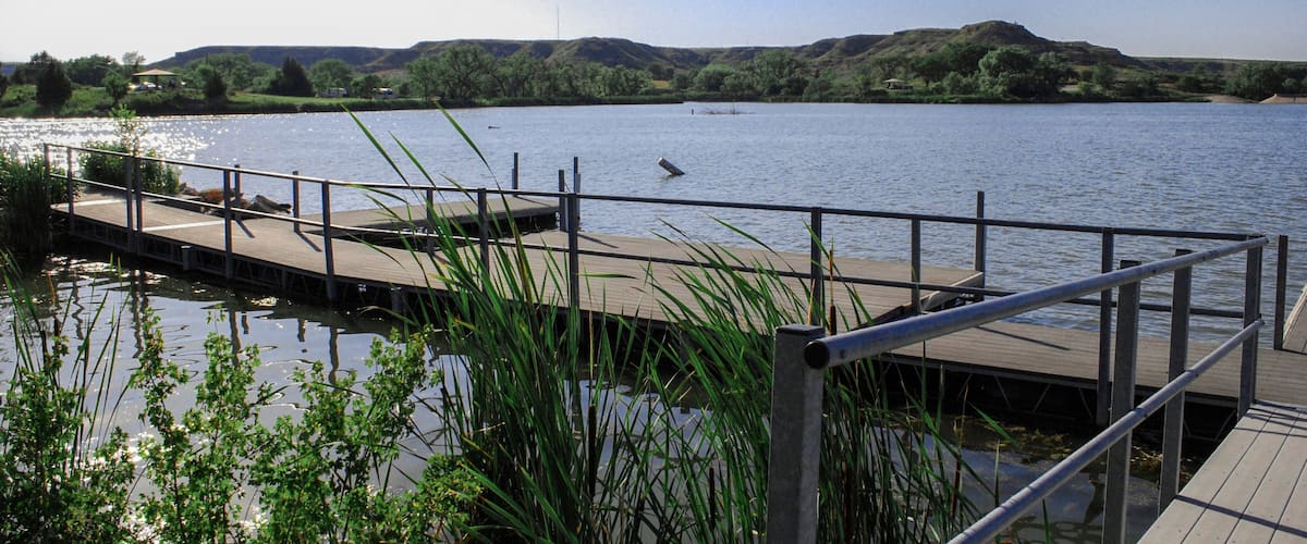 A wooden dock extending into a calm lake, surrounded by greenery and hills in the background.